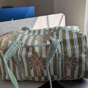 May include: A quilted travel bag with a green and white striped pattern and palm tree motifs. The bag has green straps and trim. The bag is sitting on a desk near a window.