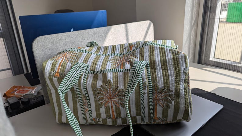 May include: A quilted travel bag with a green and white striped pattern and palm tree motifs. The bag has green straps and trim. The bag is sitting on a desk near a window.