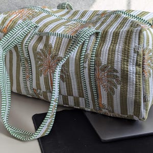 May include: A quilted duffel bag with a striped pattern in white and olive green, featuring a repeating palm tree design in orange and brown. The bag has green handles and trim.