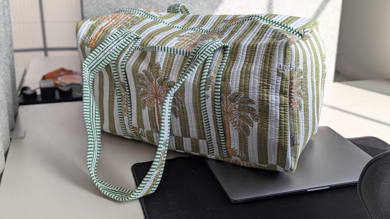 May include: A quilted duffel bag with a striped pattern in white and olive green, featuring a repeating palm tree design in orange and brown. The bag has green handles and trim.