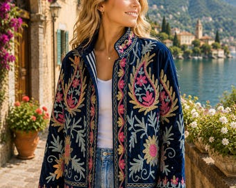 May include: A navy blue velvet jacket featuring a high collar and long sleeves, adorned with intricate floral and paisley embroidery in shades of pink, yellow, and light blue. The jacket is worn over a white top and light blue denim jeans.