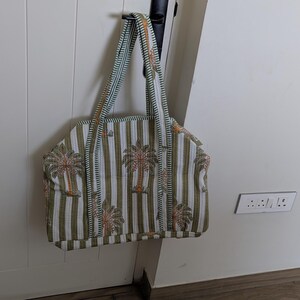 May include: A tote bag with a striped pattern in white and olive green, featuring palm tree designs. The bag has matching striped handles and is hanging on a white door.
