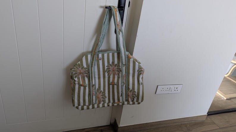 May include: A tote bag with a striped pattern in white and olive green, featuring palm tree designs. The bag has matching striped handles and is hanging on a white door.