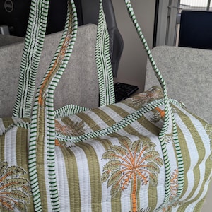 May include: Quilted duffel bag with a green and white striped pattern and palm tree motifs. The bag has green striped handles and trim. The bag is made of a soft, quilted fabric.