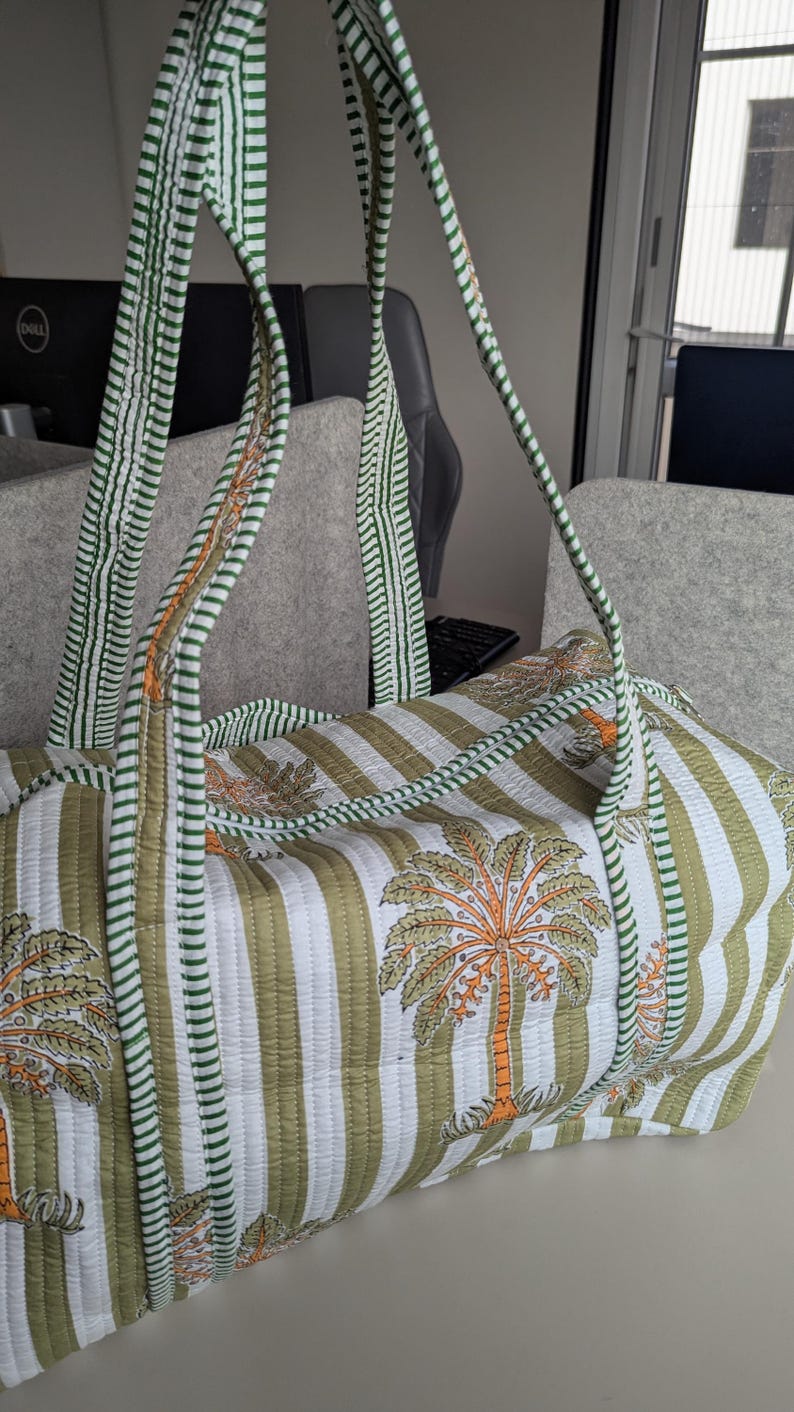 May include: Quilted duffel bag with a green and white striped pattern and palm tree motifs. The bag has green striped handles and trim. The bag is made of a soft, quilted fabric.