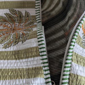 May include: Close-up of a quilted bag with a white zipper. The bag features a striped pattern in white and olive green, with a floral design in orange and green. The interior lining has a striped pattern.