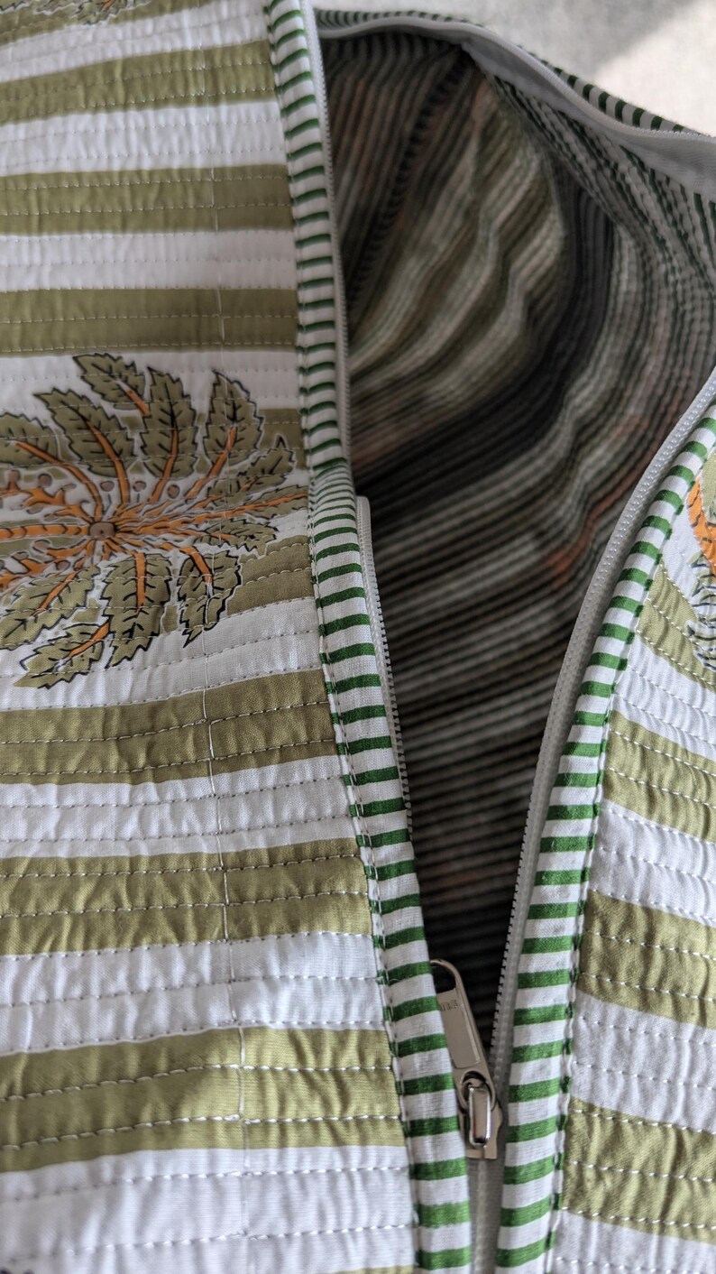 May include: Close-up of a quilted bag with a white zipper. The bag features a striped pattern in white and olive green, with a floral design in orange and green. The interior lining has a striped pattern.