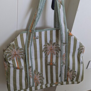 May include: Quilted tote bag with a striped pattern in olive green and white. The bag features a repeating design of palm trees in orange and brown. The handles and trim are green with a striped pattern. The bag is hanging on a black door handle.