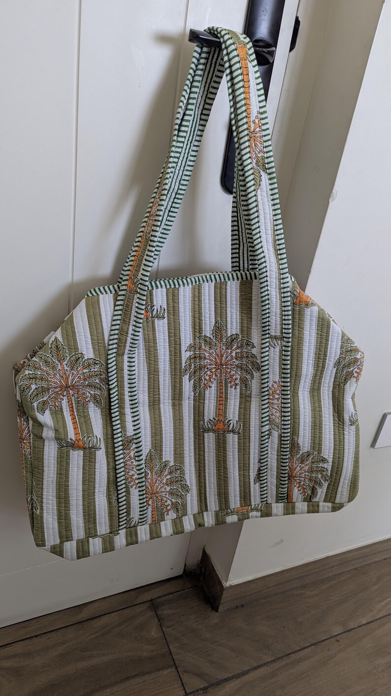 May include: Quilted tote bag with a striped pattern in olive green and white. The bag features a repeating design of palm trees in orange and brown. The handles and trim are green with a striped pattern. The bag is hanging on a black door handle.