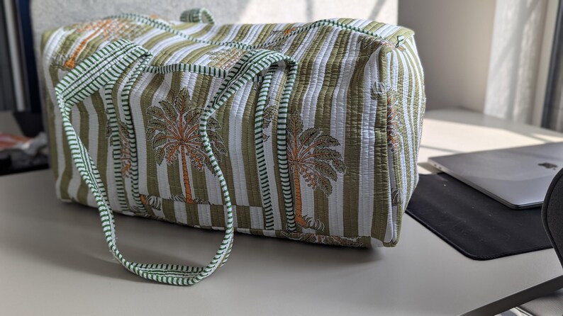 May include: A quilted tote bag with a green and white striped pattern and palm tree motifs. The bag has green handles and trim. The bag is rectangular and appears to be made of a soft fabric.