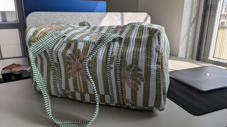 May include: A quilted travel bag with a striped pattern in white, green, and olive. The bag features a palm tree design and green handles. The bag is sitting on a white surface.