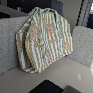 May include: A striped tote bag with a palm tree design. The bag features vertical stripes in olive green and white, with orange and brown palm tree illustrations. It has two handles and a pocket on the side.