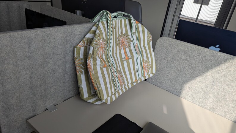 May include: A striped tote bag with a palm tree design. The bag features vertical stripes in olive green and white, with orange and brown palm tree illustrations. It has two handles and a pocket on the side.