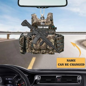 May include: A car hanging ornament shaped like a camouflage tactical vest with a black rifle attached. The ornament has the text "YOUR NAME" and a small American flag. The text "NAME CAN BE CHANGED" is also visible.