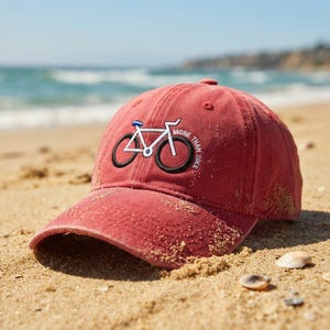 May include: A red baseball cap featuring an embroidered graphic of a bicycle in white and black thread. The text MORE THAN BIKE is stitched in a circular pattern around the bicycle graphic. The cap has a curved brim and a structured crown.