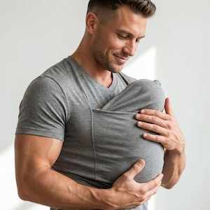May include: A gray V-neck shirt with a built-in pouch. The shirt is made of a soft material and is designed to hold a baby. The person in the image is holding the baby in the pouch.