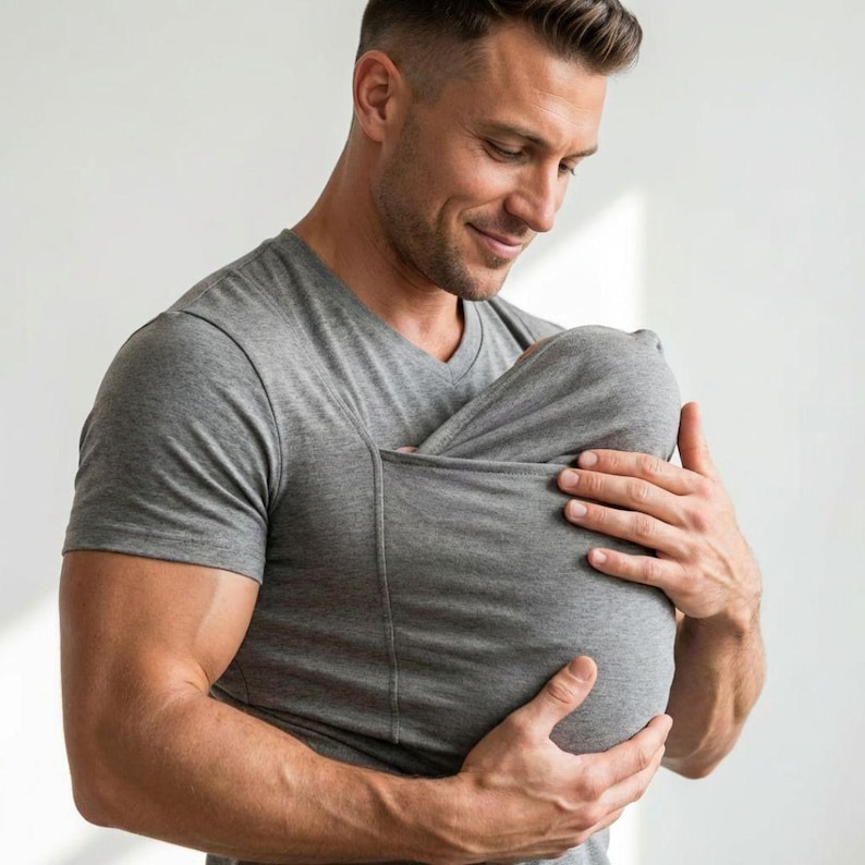 May include: A gray V-neck shirt with a built-in pouch. The shirt is made of a soft material and is designed to hold a baby. The person in the image is holding the baby in the pouch.
