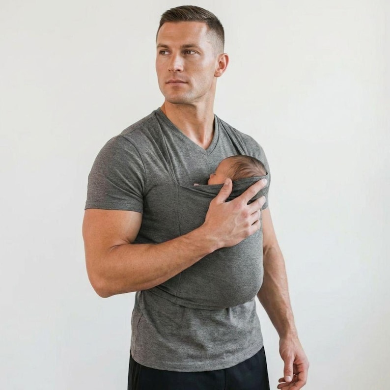 May include: A gray V-neck shirt with a built-in pouch designed to hold a baby. The shirt is made of a soft material and is being worn by a person. The baby is nestled in the pouch, with its head visible.