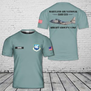 May include: A sage green short-sleeve t-shirt featuring a front graphic with the name DAVID, a 135th Airlift Group emblem, and a small US flag. The back displays a C-130J aircraft graphic with the text MARYLAND AIR NATIONAL GUARD 135TH AIRLIFT GROUP C-130J.