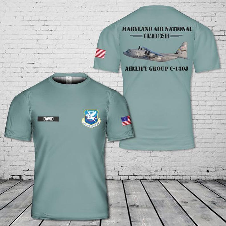 May include: A sage green short-sleeve t-shirt featuring a front graphic with the name DAVID, a 135th Airlift Group emblem, and a small US flag. The back displays a C-130J aircraft graphic with the text MARYLAND AIR NATIONAL GUARD 135TH AIRLIFT GROUP C-130J.