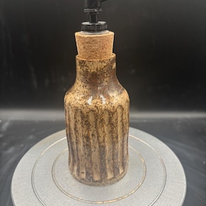 May include: A ceramic soap dispenser with a brown and tan speckled finish. The bottle has a textured, vertical ribbed design and is topped with a cork and a black pump. The dispenser is designed for liquid soap.