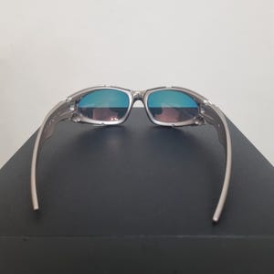 May include: Silver-toned sunglasses featuring a wrap-around frame design with metallic temples and hinges. The lenses exhibit a reflective blue and purple gradient tint, providing a distinct color shift across the surface of the eyewear.