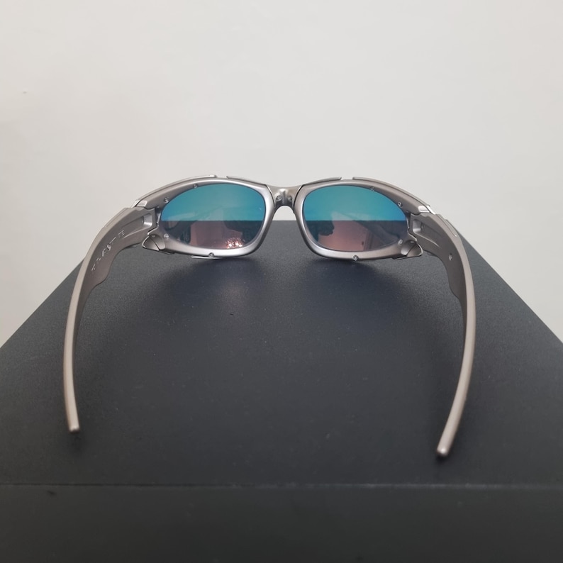 May include: Silver-toned sunglasses featuring a wrap-around frame design with metallic temples and hinges. The lenses exhibit a reflective blue and purple gradient tint, providing a distinct color shift across the surface of the eyewear.