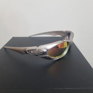 May include: Silver-toned sunglasses featuring a wrap-around frame design with metallic accents and a prominent logo on the temple. The lenses have a reflective, warm-toned orange and yellow gradient finish, set within a structured, aerodynamic frame.