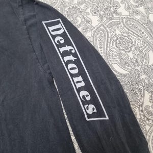 May include: Dark gray long-sleeve shirt with the word "Deftones" printed in white on the sleeve. The shirt has a faded, vintage look. The text is in a rectangular white box.