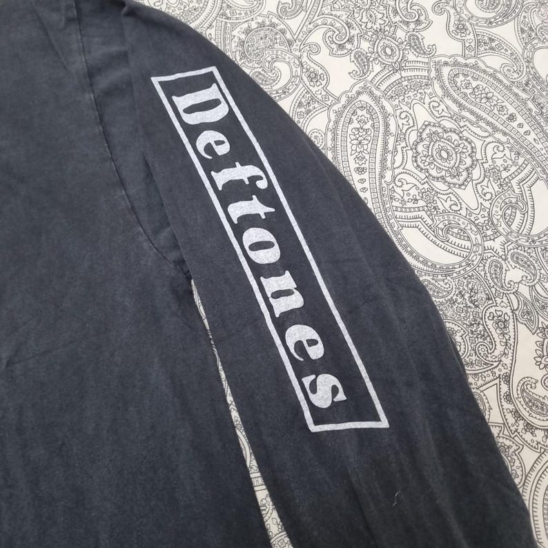 May include: Dark gray long-sleeve shirt with the word "Deftones" printed in white on the sleeve. The shirt has a faded, vintage look. The text is in a rectangular white box.