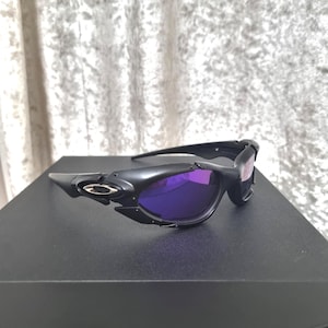 Peut inclure: Lunettes de soleil noires avec verres violets. Les lunettes ont un design élégant et moderne avec une monture noire mate et un accent argenté sur le côté. Les verres sont de couleur violette vibrante, offrant un look élégant.