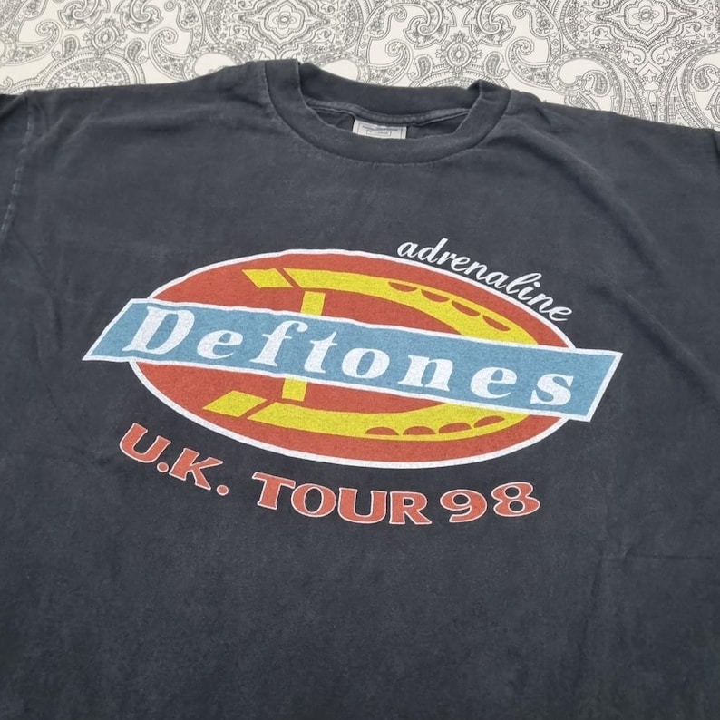 May include: Dark gray t-shirt featuring the band name "Deftones" in a blue and white oval design with the word "adrenaline" above. The text "U.K. TOUR 98" is printed below in red. A vintage band tee.