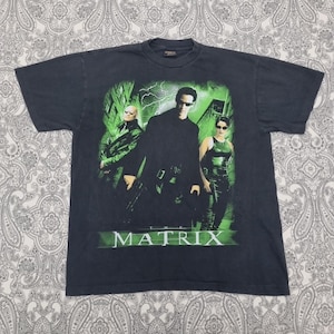 May include: Black t-shirt featuring a graphic from the movie "The Matrix." The design includes three characters in sunglasses, with a green and black color scheme. The movie title "MATRIX" is printed in green.