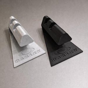 May include: Two Oakley branded accessories. One is silver, the other is black. Each has a triangular base with the Oakley logo and a curved top with three grooves. The accessories are designed to hold something.