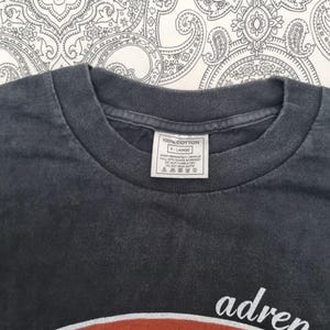 May include: Close-up of a dark gray t-shirt with a crew neck. The shirt has a white tag that reads "100% COTTON" and "X-LARGE". The bottom of the shirt has the word "adren" in white.