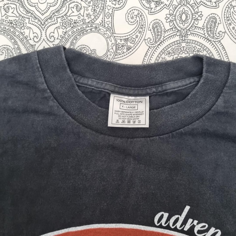 May include: Close-up of a dark gray t-shirt with a crew neck. The shirt has a white tag that reads "100% COTTON" and "X-LARGE". The bottom of the shirt has the word "adren" in white.