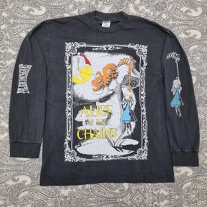 May include: Black long-sleeve t-shirt featuring a graphic from the band Alice in Chains. The design includes illustrations of characters and the band's name in yellow. The left sleeve has the band's name in a vertical design.