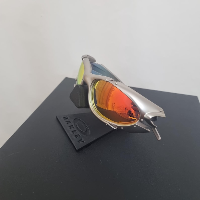 May include: Silver-toned sunglasses with a metallic frame and reflective orange-tinted lenses. The eyewear is mounted on a black display stand featuring the embossed Oakley logo. The design includes a wrap-around frame structure with visible rivets.