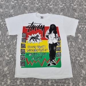 May include: White t-shirt with a graphic design featuring the Stüssy logo in red, a lion, and a person. The design includes text in yellow and green. The shirt has short sleeves and a crew neck.