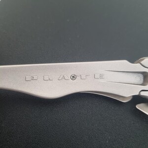 May include: A silver-toned metal multi-tool handle featuring a series of engraved symbols and letters including squares, a stylized letter A, a cross symbol, and the letter T. The handle has a smooth, matte finish with a curved, ergonomic shape.