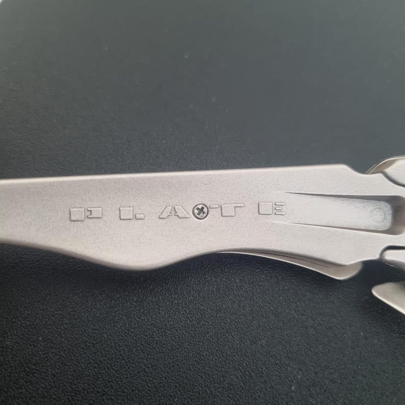 May include: A silver-toned metal multi-tool handle featuring a series of engraved symbols and letters including squares, a stylized letter A, a cross symbol, and the letter T. The handle has a smooth, matte finish with a curved, ergonomic shape.