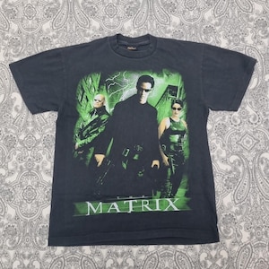 May include: Dark gray t-shirt featuring a graphic from the movie "The Matrix." The design includes images of three characters in front of a green cityscape with the film's title in green letters.