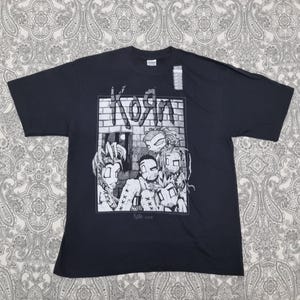 May include: Black t-shirt featuring a graphic of the band Korn with the band's name in a graffiti-style font. The graphic is in black and white and includes cartoon-style illustrations of the band members. The website address korn.com is also visible.
