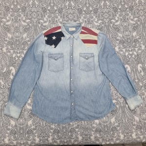 Może przedstawiać: Jasnoniebieska koszula dżinsowa z długimi rękawami i zapięciem na guziki. Koszula posiada dwie kieszenie na piersi i wzór amerykańskiej flagi na ramionach. Wzór flagi zawiera czerwono-białe paski i niebieskie pole z białymi gwiazdami.