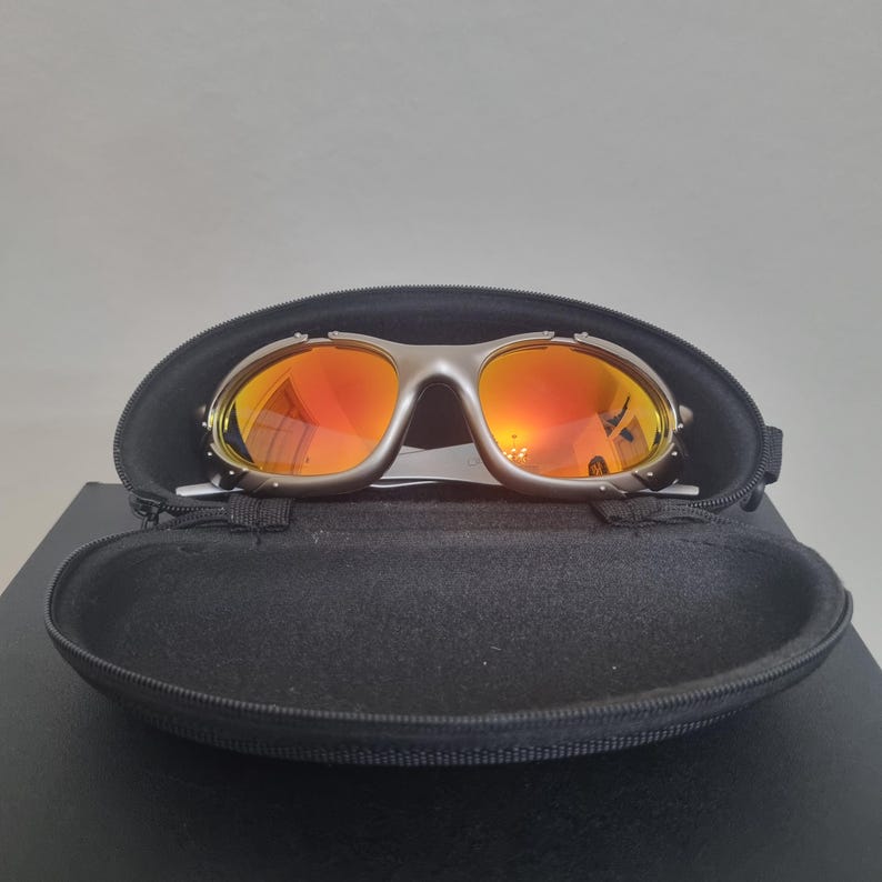 May include: Silver-toned sunglasses with reflective orange lenses featuring a wrap-around frame design. The sunglasses are positioned inside a black, oval-shaped, zippered hard-shell protective case with a soft interior lining.