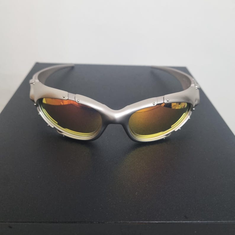 May include: Wraparound sunglasses featuring a matte silver metallic frame and reflective orange-tinted lenses. The design includes a segmented frame structure with visible rivets along the temples and bridge, creating a technical, industrial aesthetic.