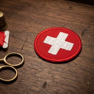 May include: A red and white Swiss flag patch, a gold thimble, red thread, and small gold scissors on a wooden surface. The patch features a white cross on a red background, and the sewing tools suggest a crafting theme.