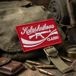 May include: A red patch with white embroidered text reading "Kalashnikova CLASSIC" and a rifle graphic. The patch is set against a backdrop of military-style gear, including a canvas bag, bullets, and a compass.