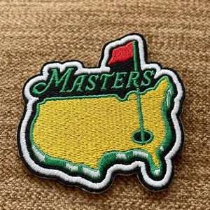May include: Embroidered patch featuring the Masters Tournament logo. The patch is shaped like a map of the United States, primarily yellow with green and white borders. The word "MASTERS" is in green script, and a red flag is visible.