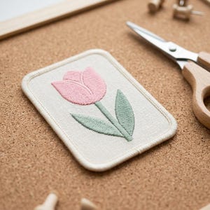 May include: A rectangular patch with a pink tulip embroidery on a light beige background. The patch is on a corkboard with wooden push pins and scissors with wooden handles.